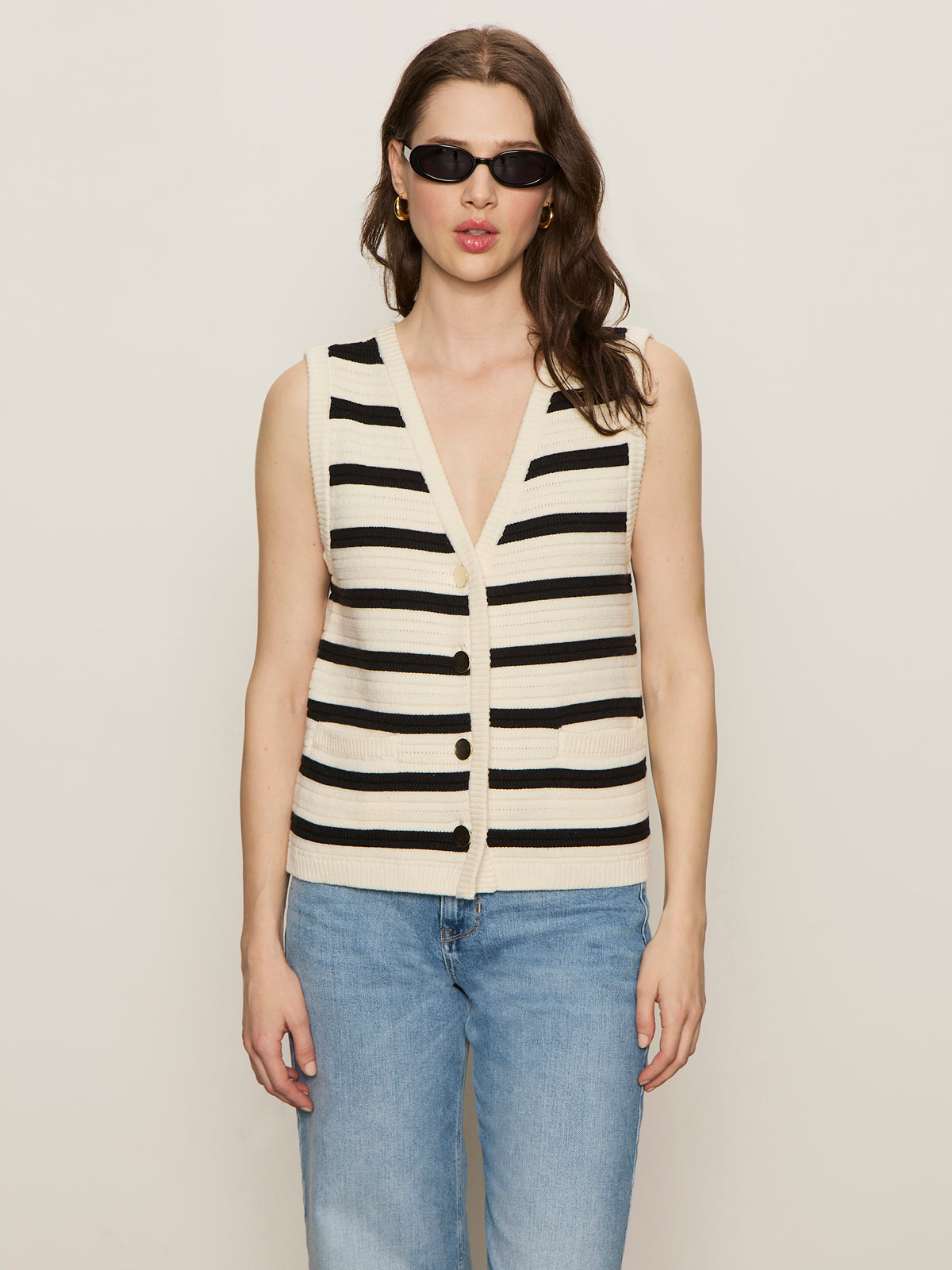 A woman wears the Sanctuary Clothing textured sweater vest macrame black stripe 3, paired with black sunglasses and light blue jeans, standing against a plain light background.