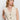 A woman with long, wavy blonde hair models the Sanctuary Clothing textured sweater vest macrame—a sleeveless, cream knit vest with black buttons and front pockets—against a plain white background, looking slightly to the side.