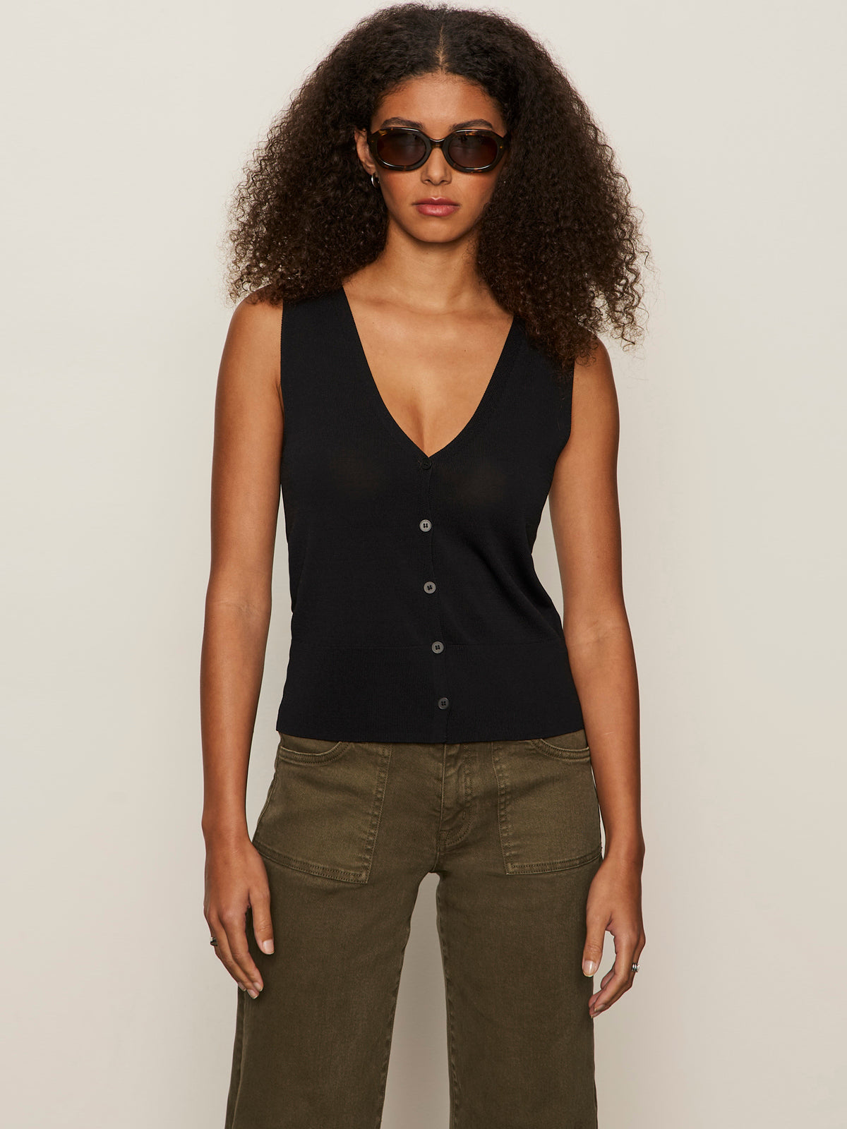 A woman with curly hair wears dark sunglasses and the Sanctuary Clothing knitted button front black top, paired with olive green pants, standing against a plain light background.