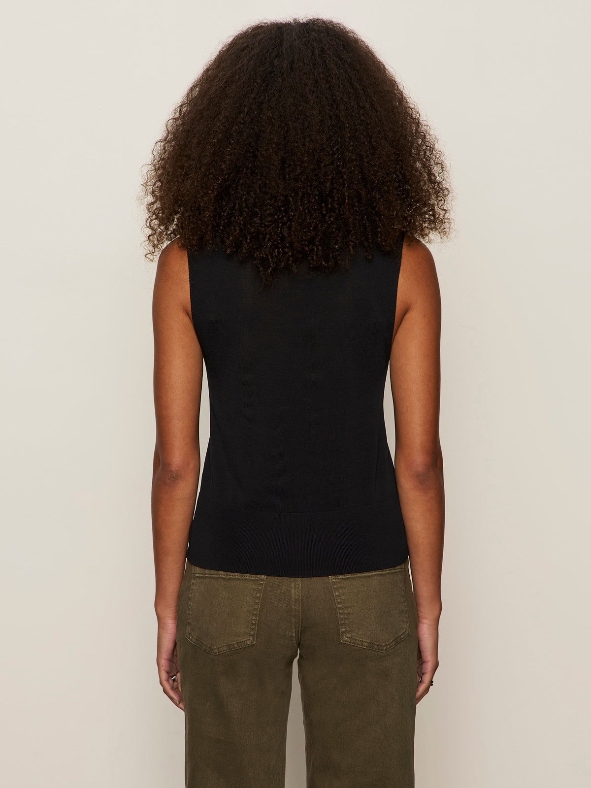 A person with curly hair is seen from behind, wearing the Sanctuary Clothing knitted button front black top and olive green pants, standing against a plain light background.