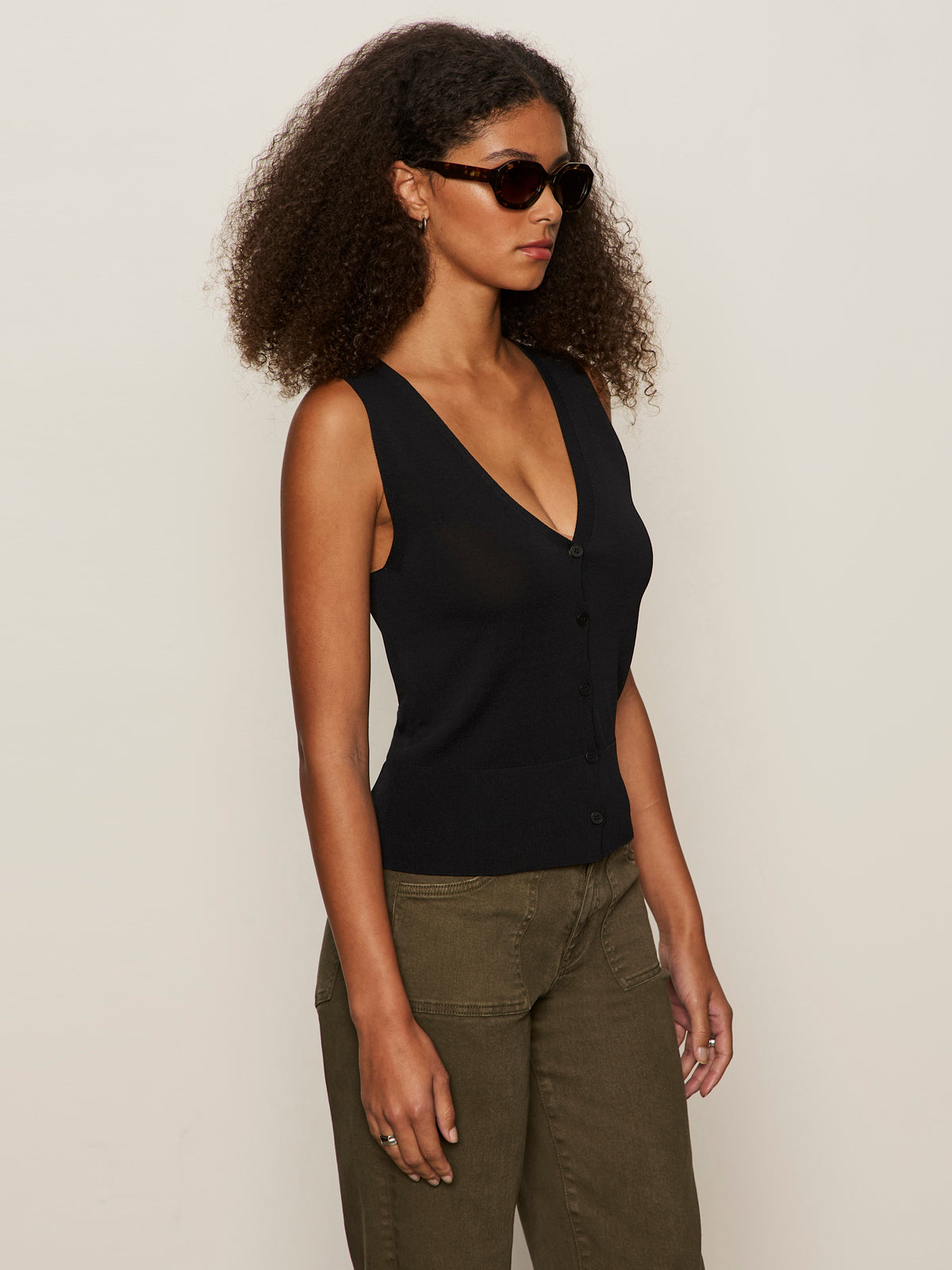 A woman with curly hair wears sunglasses and the Sanctuary Clothing knitted button front black top, paired with olive-green pants, standing against a plain light background, facing slightly to the side.