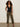A woman with curly hair wears sunglasses and the Sanctuary Clothing knitted button front black top with olive green wide-leg pants and black sandals, standing against a plain light-colored wall.