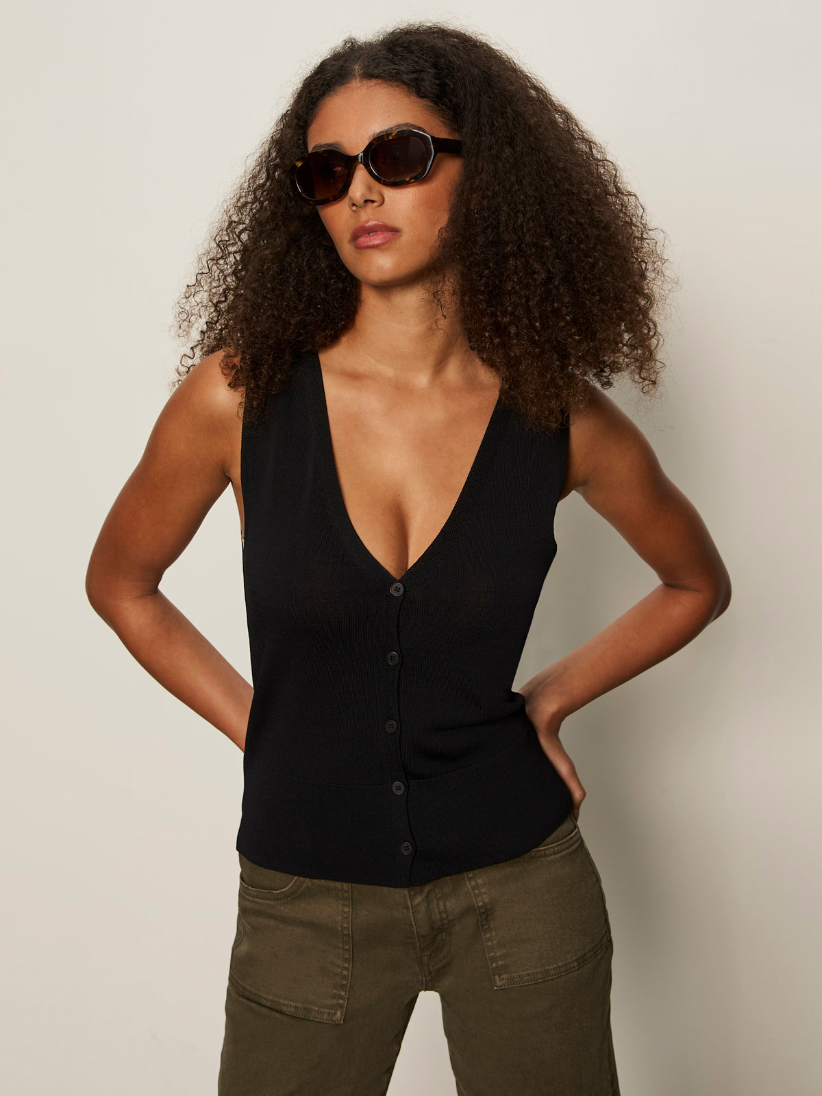 A woman with curly hair and dark sunglasses wears the Sanctuary Clothing knitted button front black top with olive green pants, standing against a plain light background, hands on her hips.