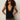 A woman with curly hair and dark sunglasses wears the Sanctuary Clothing knitted button front black top with olive green pants, standing against a plain light background, hands on her hips.