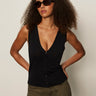 A woman with curly hair and dark sunglasses wears the Sanctuary Clothing knitted button front black top with olive green pants, standing against a plain light background, hands on her hips.
