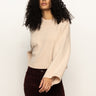 A woman with curly hair models the Sanctuary Clothing Forever Cashmere Popover in toasted beige, featuring wide sleeves, paired with burgundy pants against a plain white background. She faces forward with one arm slightly extended.