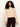 A woman with curly hair wears the Sanctuary Clothing Forever Cashmere Popover in toasted beige, paired with dark brown pants, as she stands against a plain white background and looks at the camera with a slight smile.