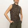 A person with light hair tied back wears the fuzzy shell french kitty sleeveless, high-neck leopard print top from Sanctuary Clothing and olive green pants, standing against a plain white background with hands in pockets.