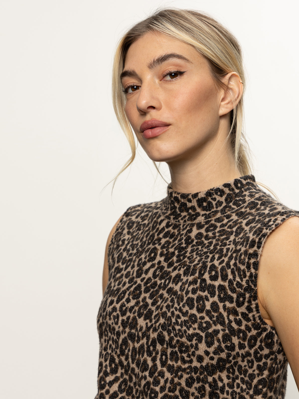 A woman with long blonde hair in a loose ponytail models the “fuzzy shell french kitty” sleeveless, high-neck top by Sanctuary Clothing. Posing against a plain light background, she gazes confidently at the camera with a neutral expression.