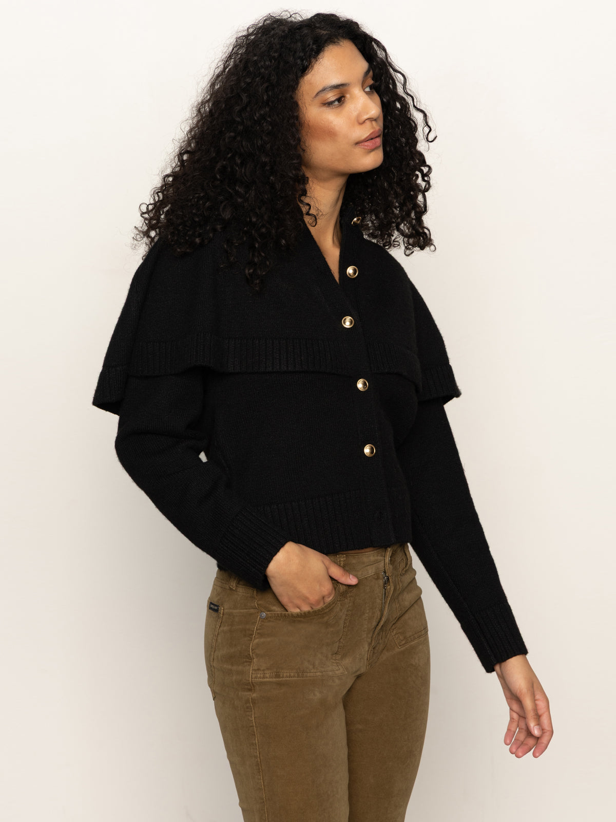 A woman with curly hair wears the Sanctuary Clothing cape cardi black sweater and brown pants, standing against a plain light background and gazing slightly to the side with a neutral expression.