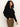 A woman with curly hair wears the Sanctuary Clothing cape cardi black sweater and brown pants, standing against a plain light background and gazing slightly to the side with a neutral expression.