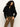 A woman with curly dark hair models the Sanctuary Clothing cape cardi black sweater, featuring a wide collar, paired with brown pants against a plain light background.