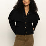 A woman with curly hair wears the Cape Cardi Black sweater by Sanctuary Clothing and brown pants. She stands against a light background with her hands in her pockets, gazing slightly to the side.