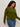 A woman with curly hair wears the Sanctuary Clothing must have cardi in martini olive and blue jeans, looking down and to the side while standing against a plain light background.