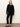 A person stands against a light wall wearing the Sanctuary Clothing must have cardi black, paired with black pants and flats, arms relaxed and face neutral.