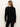 A woman with long, straight light brown hair stands with her back to the camera, wearing the Sanctuary Clothing must have cardi black and black pants against a plain light-colored wall.