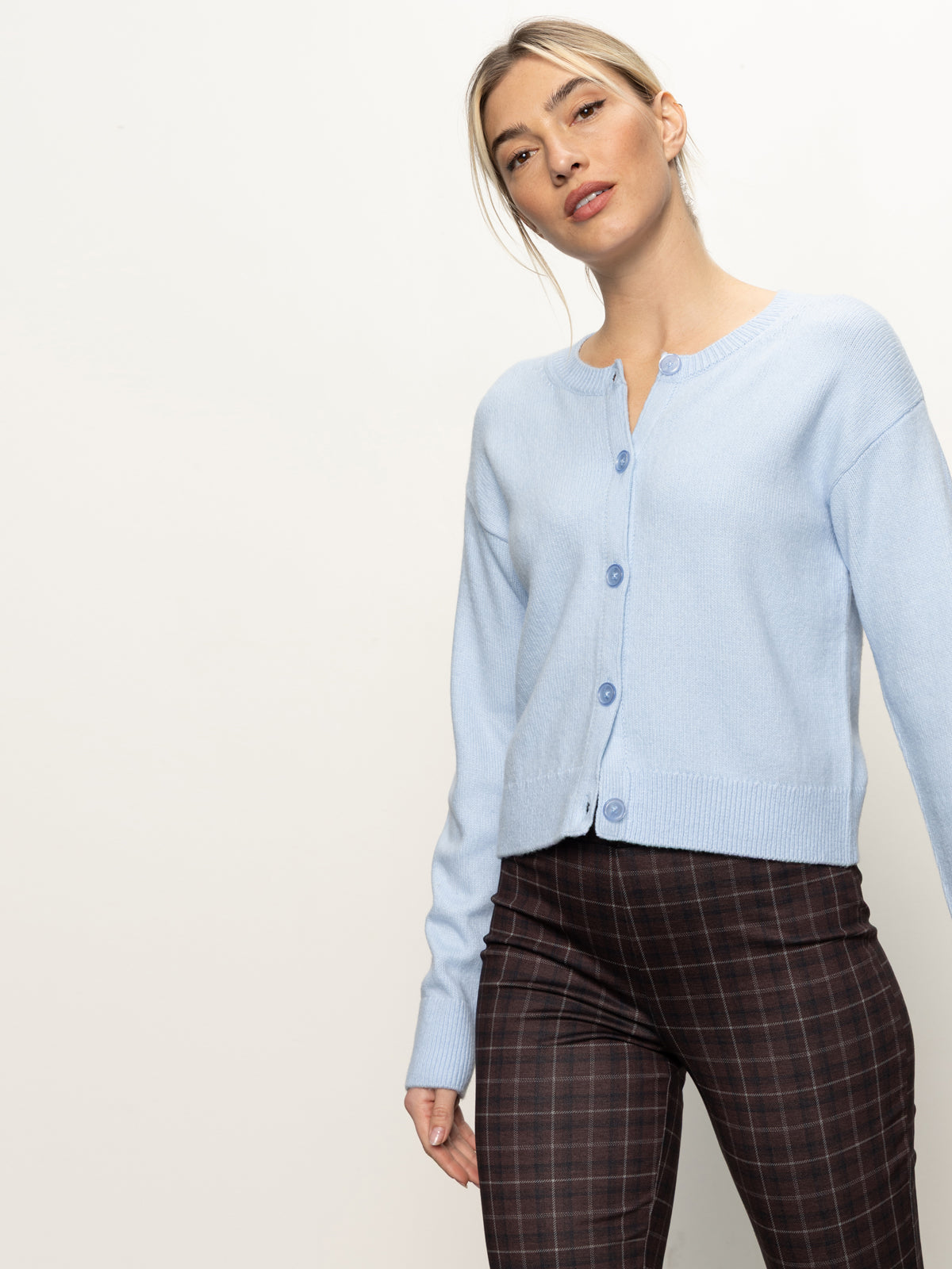 A woman wears the Sanctuary Clothing must have cardi blue powder, styled with brown plaid pants, and stands confidently against an off-white background.