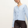 A woman wears the Sanctuary Clothing must have cardi blue powder, styled with brown plaid pants, and stands confidently against an off-white background.