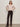 A woman stands against a plain background wearing a light pink button-up cardigan, dark brown corduroy pants with a belt, and black ankle boots. She has long brown hair and a neutral expression, with hands resting on her hips.