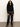 A woman with curly hair wearing the Sanctuary Clothing zip it sweater in black stands against a plain light-colored wall on a concrete floor, paired with loose shiny black pants with white side stripes.