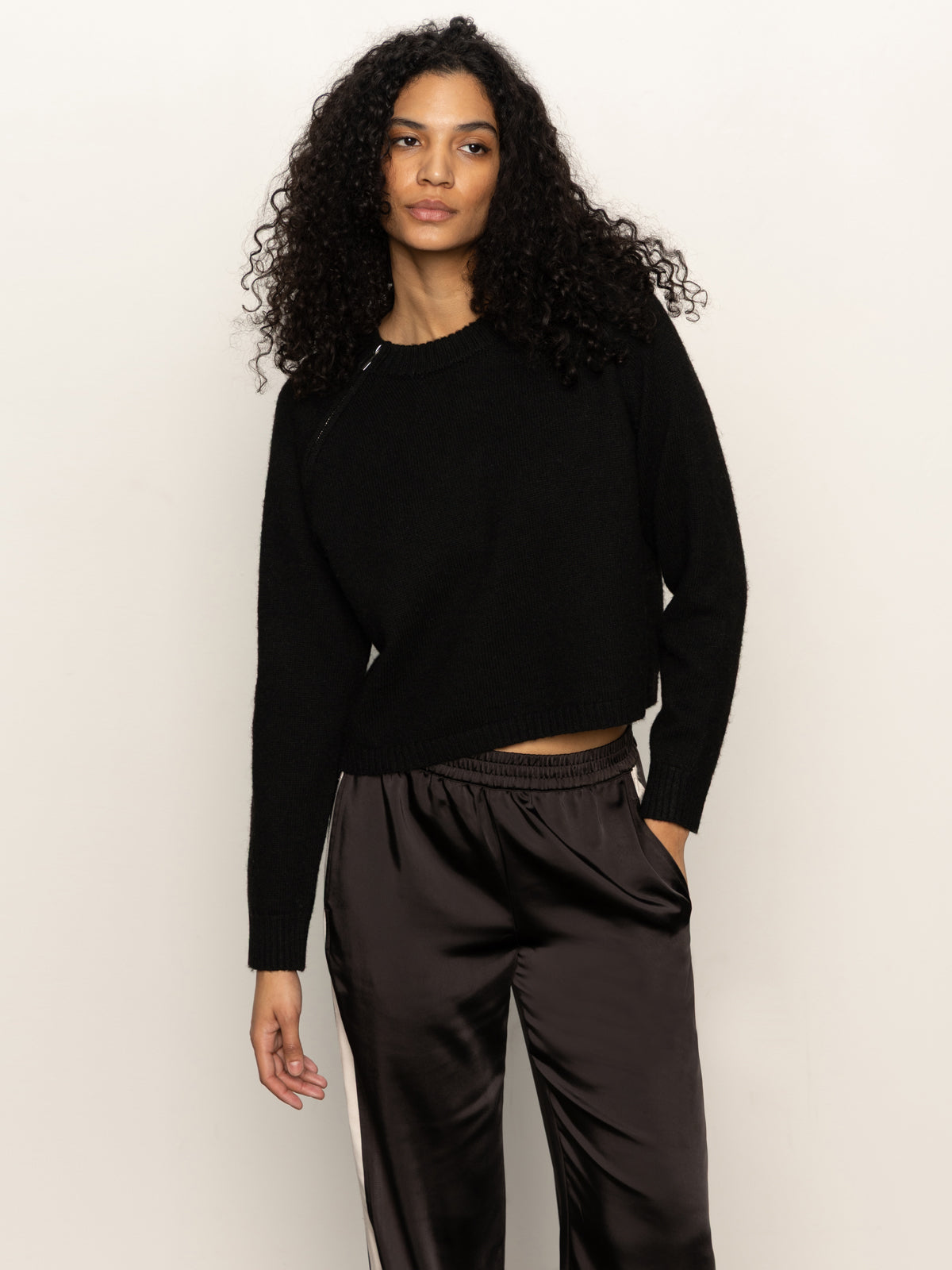 A woman with long curly hair wears the Sanctuary Clothing zip it sweater in black, paired with shiny black pants. She stands against a plain light background, one hand in her pocket, confidently looking at the camera.