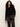 A woman with long curly hair wears the Sanctuary Clothing zip it sweater in black, paired with shiny black pants. She stands against a plain light background, one hand in her pocket, confidently looking at the camera.