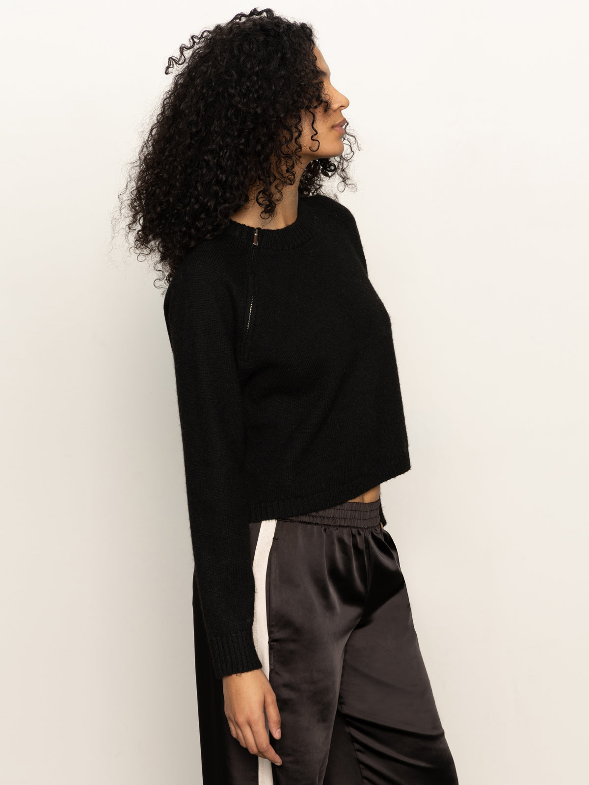 Woman with curly hair wears the Sanctuary Clothing zip it sweater in black and black satin jogger pants with white side stripes, standing sideways against a plain white background.