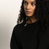 A woman with curly black hair and medium skin tone wears the Sanctuary Clothing zip it sweater in black, gazing thoughtfully to her left against a plain, light background.