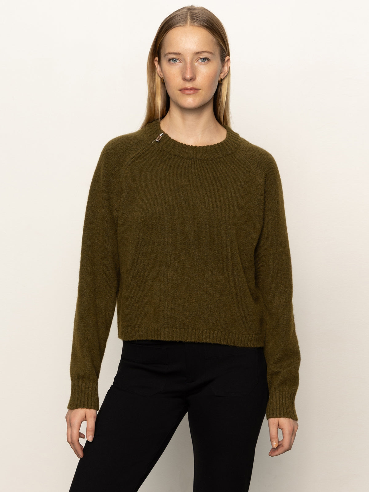 A woman with straight blonde hair wears Sanctuary Clothing's zip it sweater in dirty martini and black pants, standing against a plain off-white background, looking directly at the camera with a neutral expression.