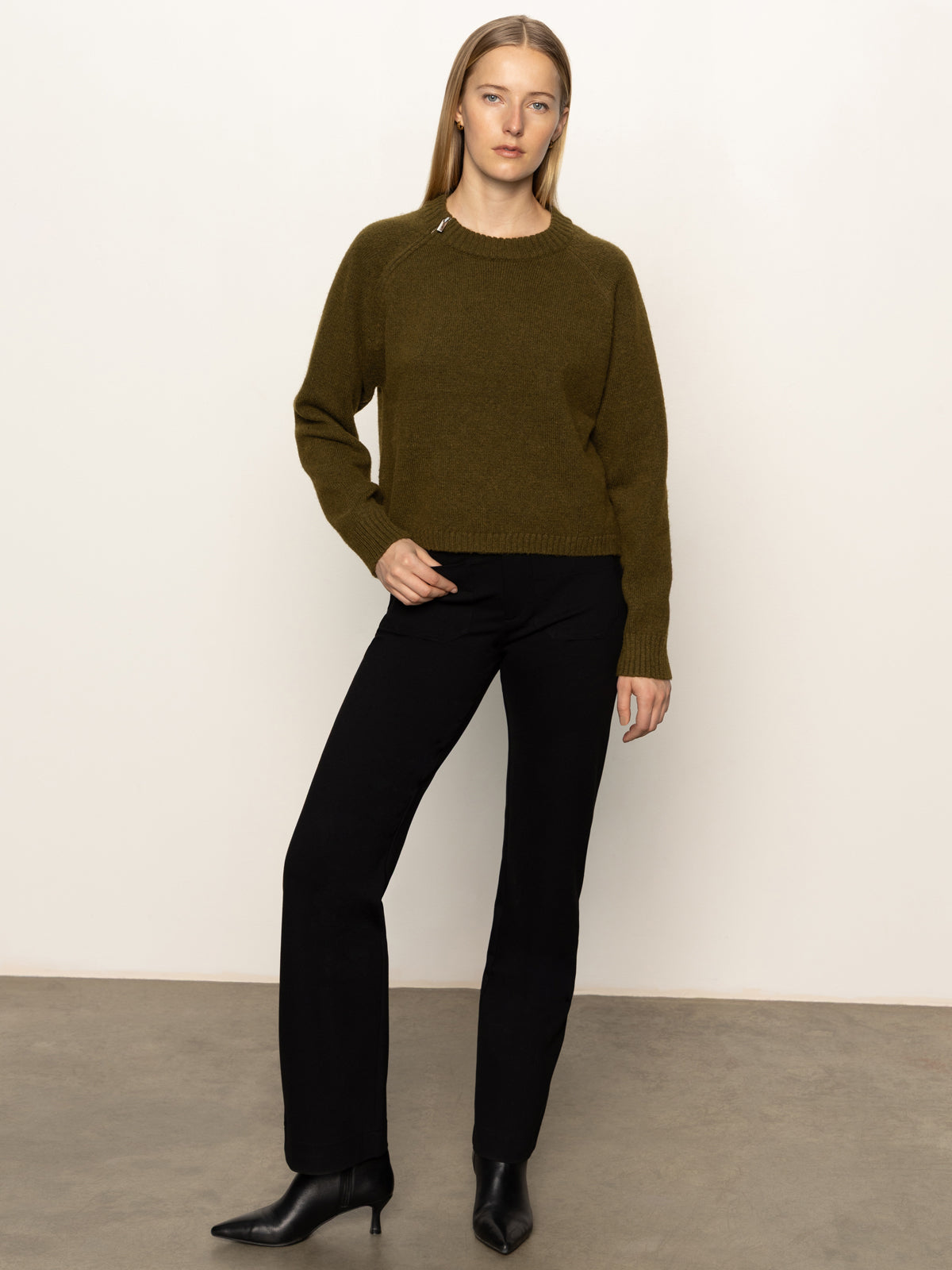 A woman with long blonde hair models the Sanctuary Clothing zip it sweater in dirty martini, paired with black pants and black heeled boots, standing against a plain white wall and concrete floor.