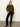 A woman with long blonde hair models the Sanctuary Clothing zip it sweater in dirty martini, paired with black pants and black heeled boots, standing against a plain white wall and concrete floor.