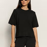 A woman with long dark hair wears black sunglasses and the perfect sweater tee in black by Sanctuary Clothing, paired with black pants, standing against a plain light background.