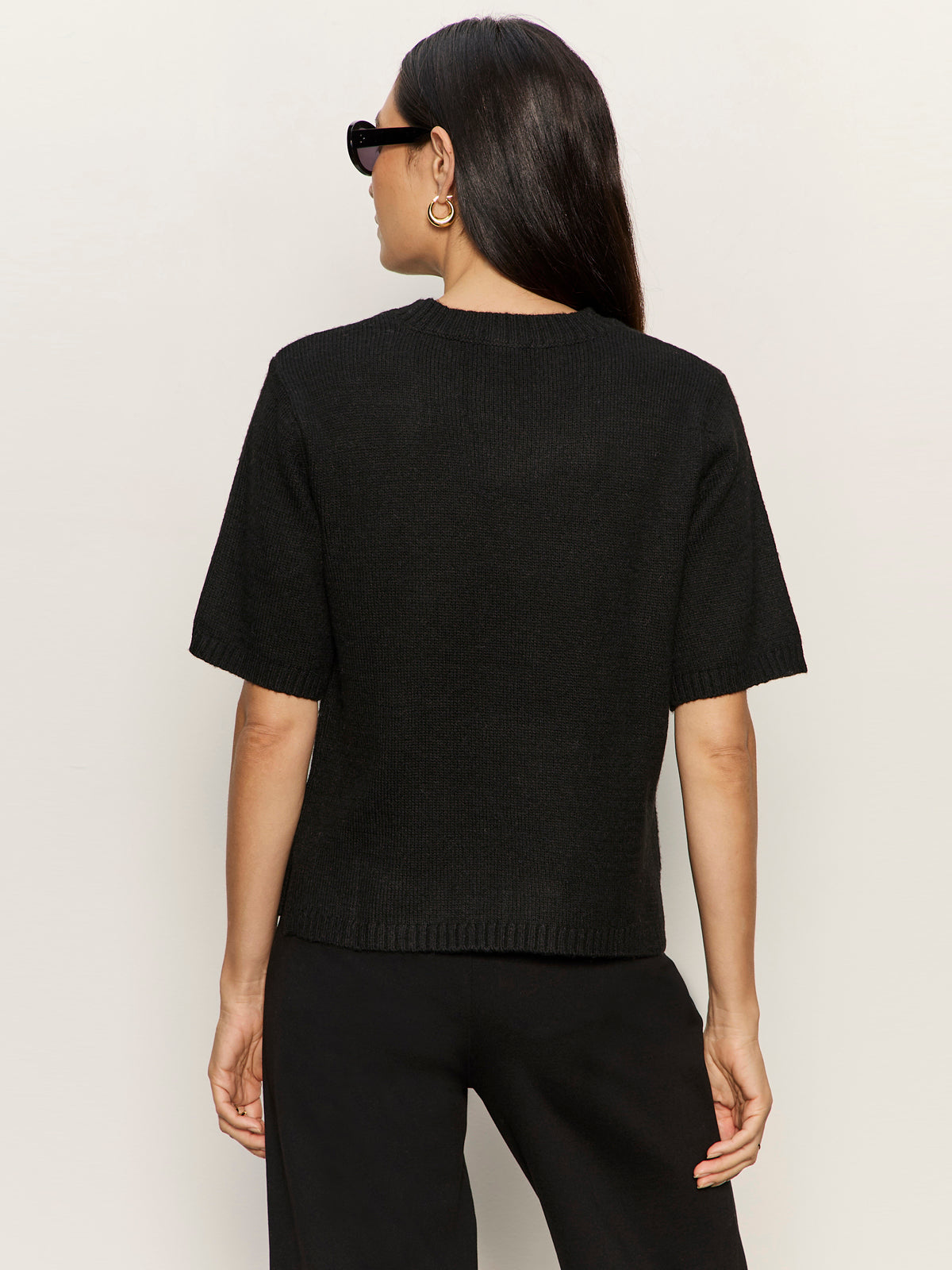 A woman with long dark hair, wearing black sunglasses, gold hoop earrings, and the Sanctuary Clothing perfect sweater tee in black with matching pants, is pictured from the back against a light background.