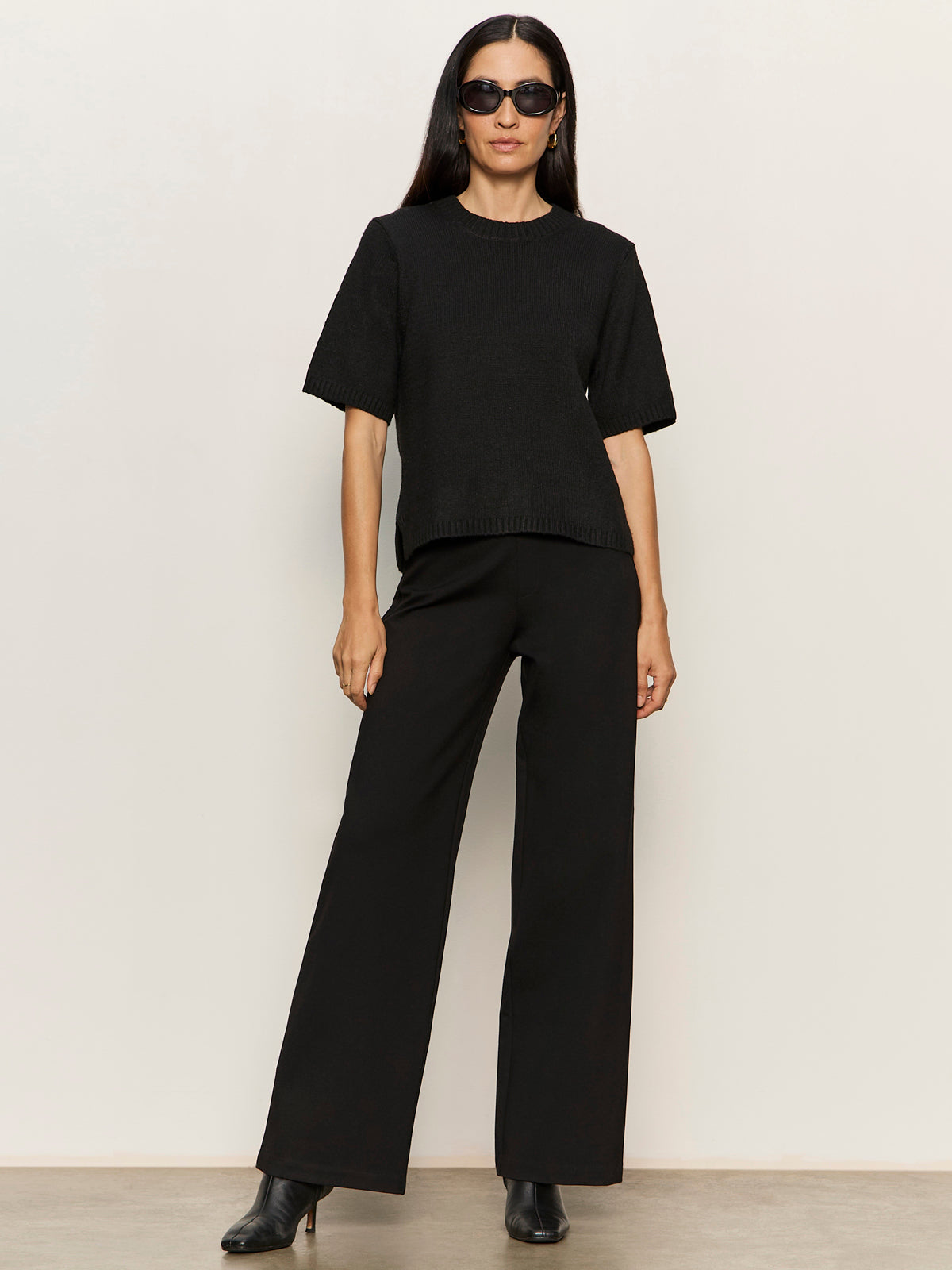 A woman stands against a plain light background in a relaxed pose, wearing Sanctuary Clothing's perfect sweater tee in black, paired with wide-leg black pants, sunglasses, and black heeled boots.