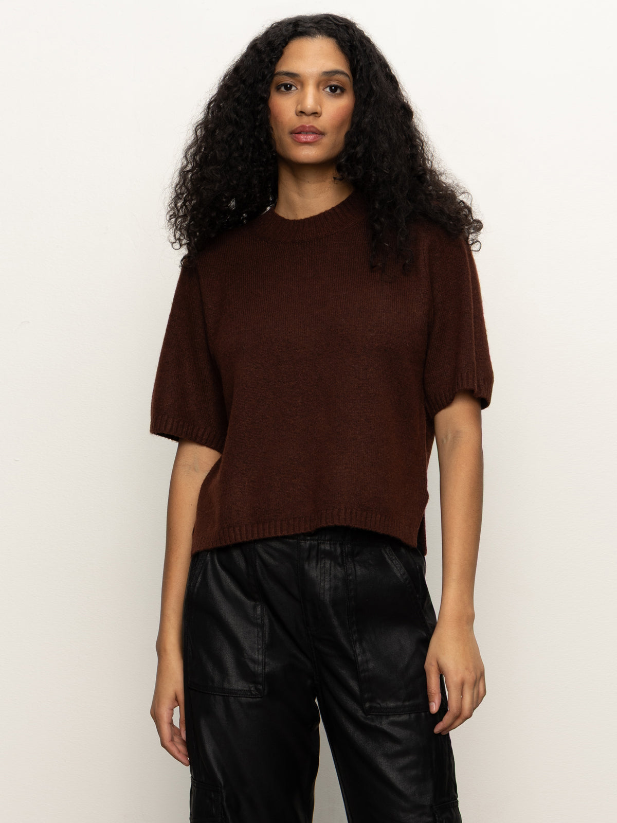 A woman with long curly hair wears the Sanctuary Clothing perfect sweater tee in chicory and black pants, standing against a plain light background.