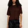 A woman with long curly hair wears the Sanctuary Clothing perfect sweater tee in chicory and black pants, standing against a plain light background.