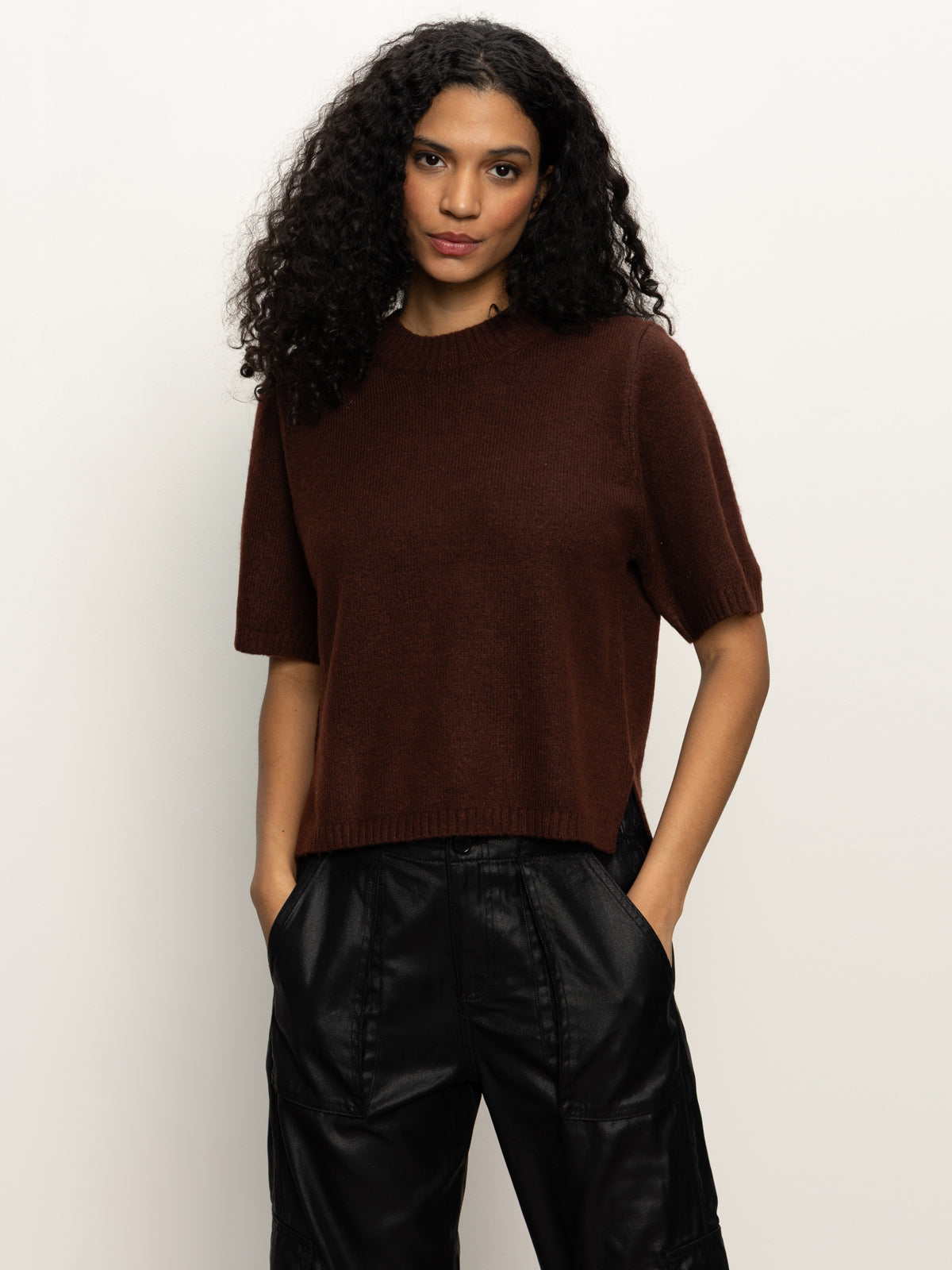 A woman with long, curly hair models the Sanctuary Clothing perfect sweater tee chicory—a short-sleeved, dark brown top—with black leather pants against a light background, hands in her pockets and looking at the camera.