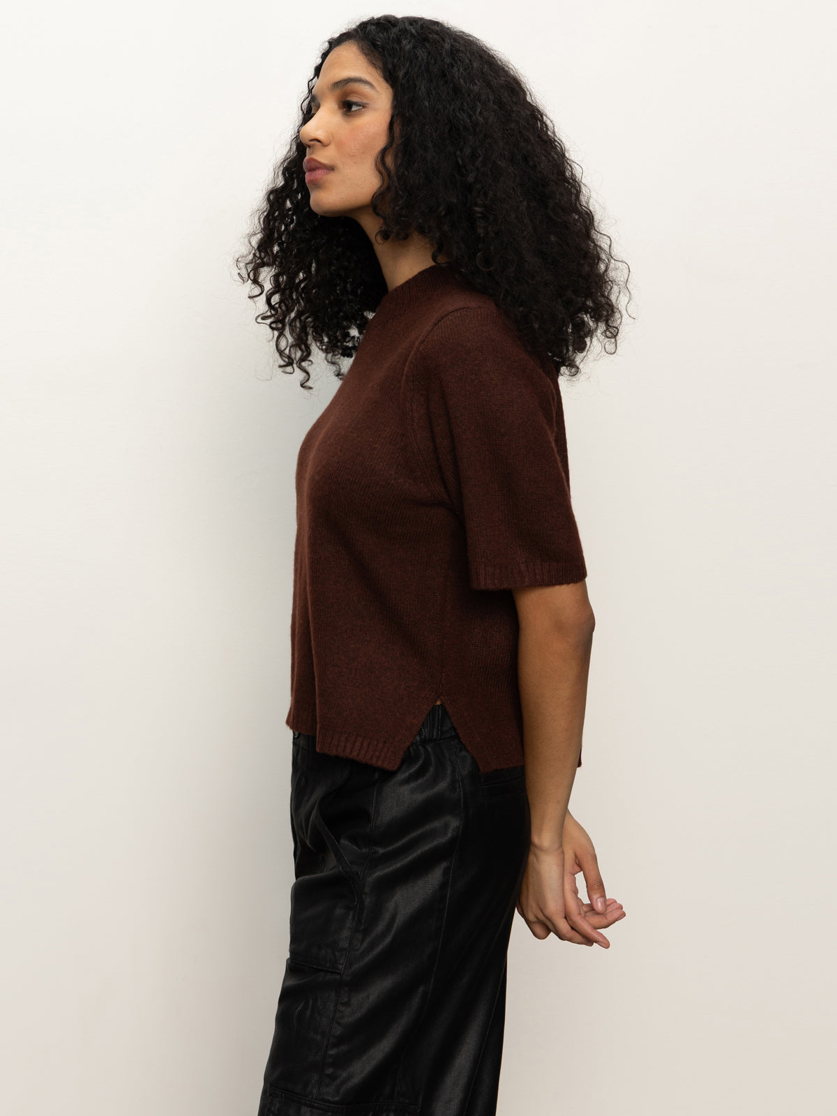 A woman with curly hair stands in profile against a plain light background, wearing the Sanctuary Clothing perfect sweater tee chicory and black pants.