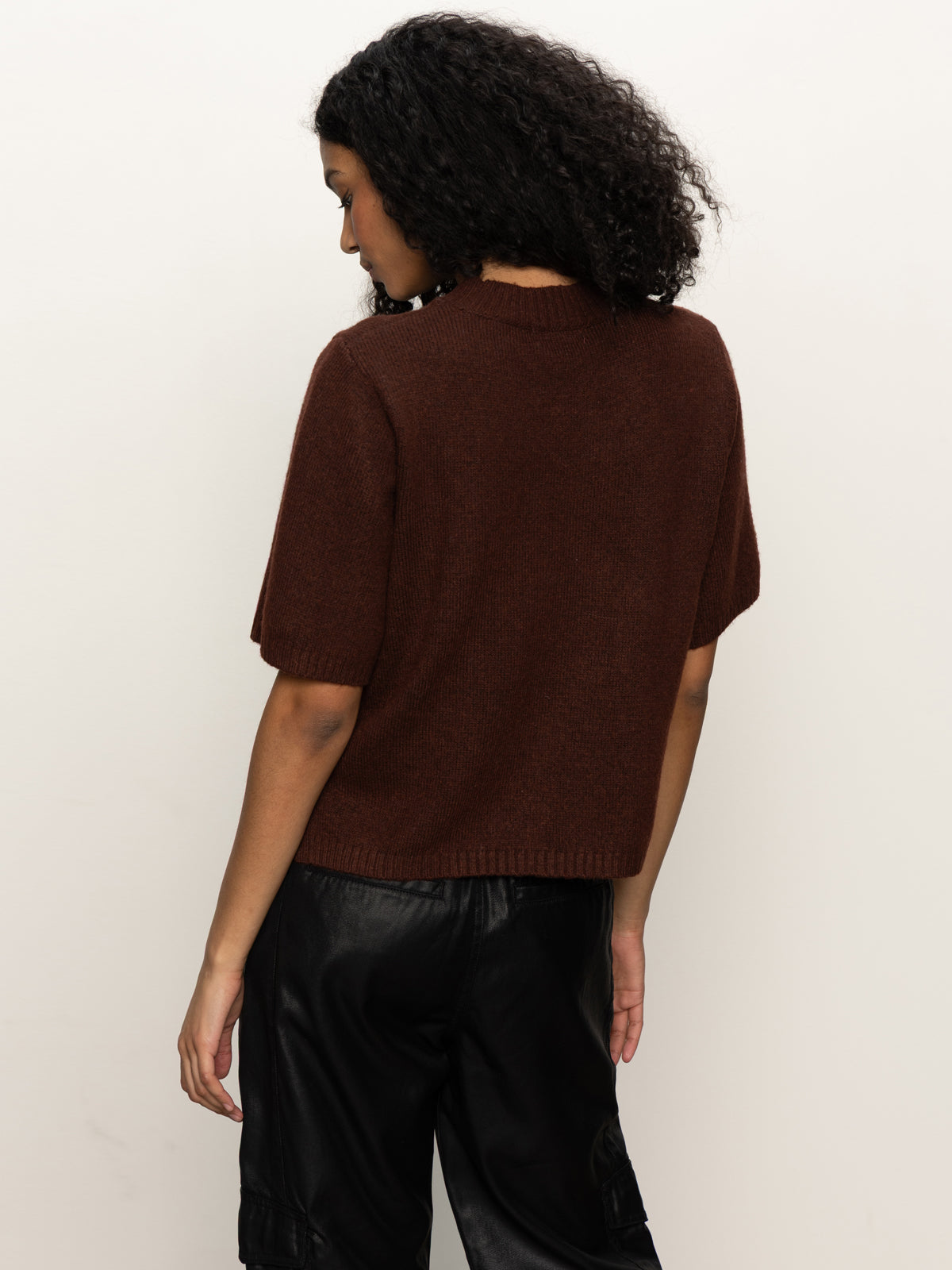 A woman with curly dark hair stands with her back to the camera, wearing Sanctuary Clothing's Perfect Sweater Tee in chicory and black pants against a plain light background.