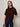 A woman with straight brown hair wears the Sanctuary Clothing Perfect Sweater Tee in dark crimson and dark blue jeans, standing against a plain light background, holding a small bag and looking at the camera.