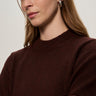 A woman with light brown hair and silver hoop earrings stands with her arms crossed, wearing the Sanctuary Clothing perfect sweater tee in dark crimson, gazing slightly to the side with a neutral expression.