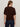 A person with long, light brown hair stands with their back to the camera, wearing Sanctuary Clothing's Perfect Sweater Tee in dark crimson and high-waisted dark blue jeans. The background is a plain, light-colored wall.