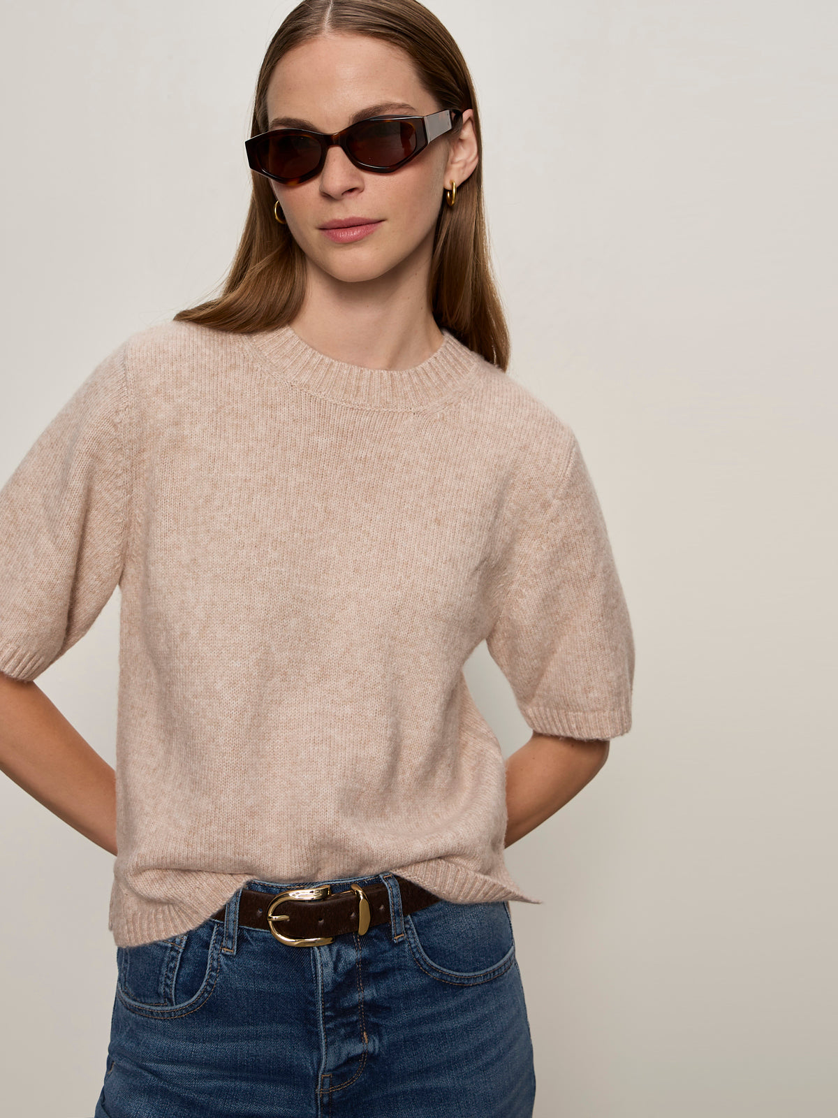 A woman with straight light brown hair and a neutral expression wears Sanctuary Clothing's perfect sweater tee fawn, dark sunglasses, blue jeans with a black belt, standing against a plain light background.