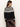 A woman with long brown hair wears the Sanctuary Clothing fairisle crewneck cardi in ink multi, paired with a dark brown satin skirt, standing with her back and side turned toward the camera against a plain background.