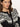 A woman with wavy brown hair wears the Sanctuary Clothing fairisle crewneck cardi in ink multi, featuring a black and white pattern and wooden buttons, as she looks seriously at the camera against a plain background.