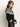 A woman with long brown hair wears the Sanctuary Clothing fairisle crewneck cardi in ink multi, featuring black and white geometric patterns. She stands in profile against a plain light background, hands loosely clasped.