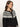A woman with wavy brown hair models the Sanctuary Clothing fairisle crewneck cardi in ink multi, featuring a white geometric pattern on the shoulders, as she gazes to the side against a plain background.