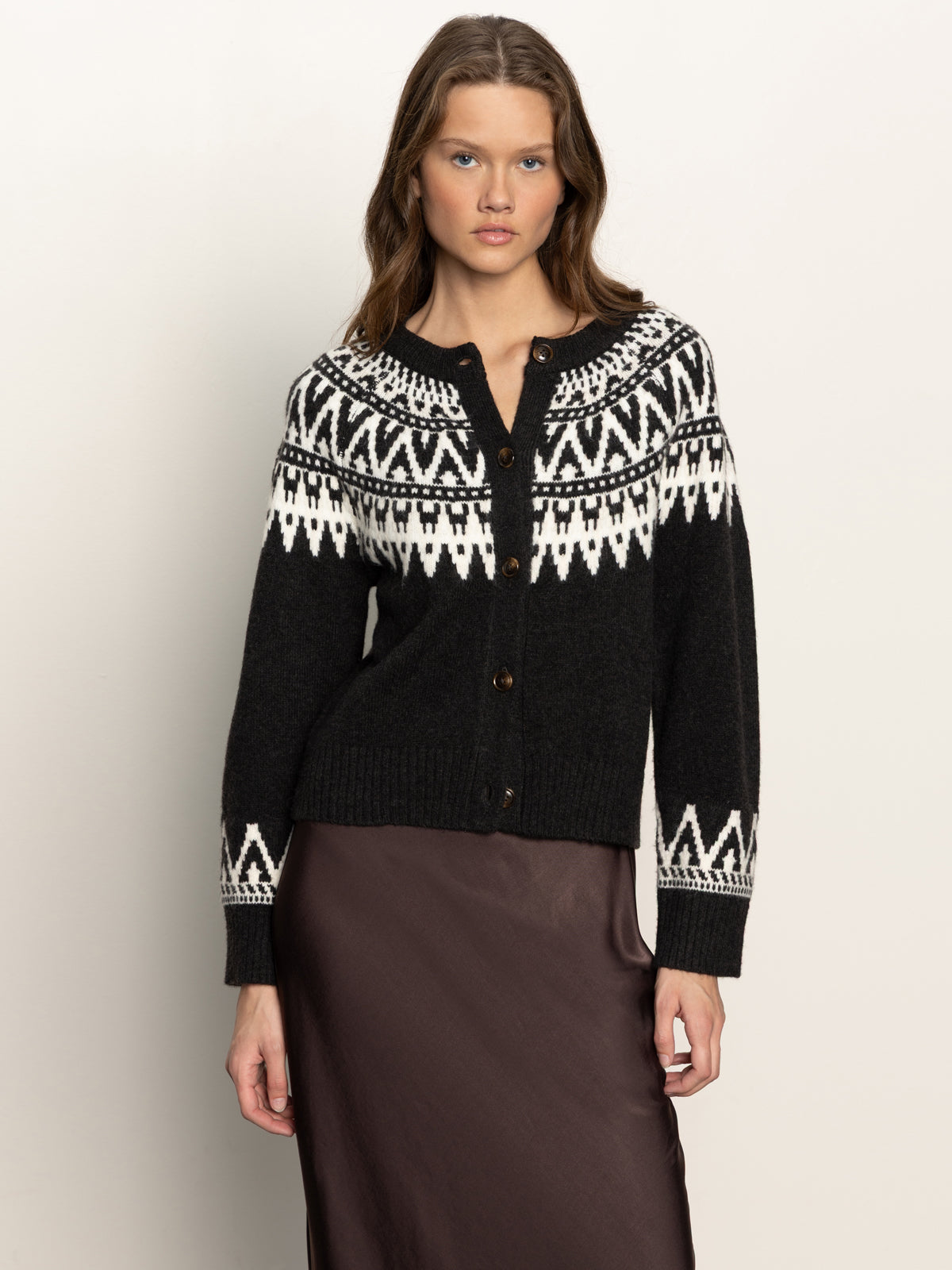 A woman wearing the Sanctuary Clothing fairisle crewneck cardi ink multi, featuring white geometric patterns, pairs it with a dark brown skirt and stands against a light background.