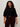 A woman with curly hair models the Sanctuary Clothing ribbed sweater tee in black (extended sizing), paired with burgundy pants and a black belt, standing against a plain light background and looking confidently at the camera.