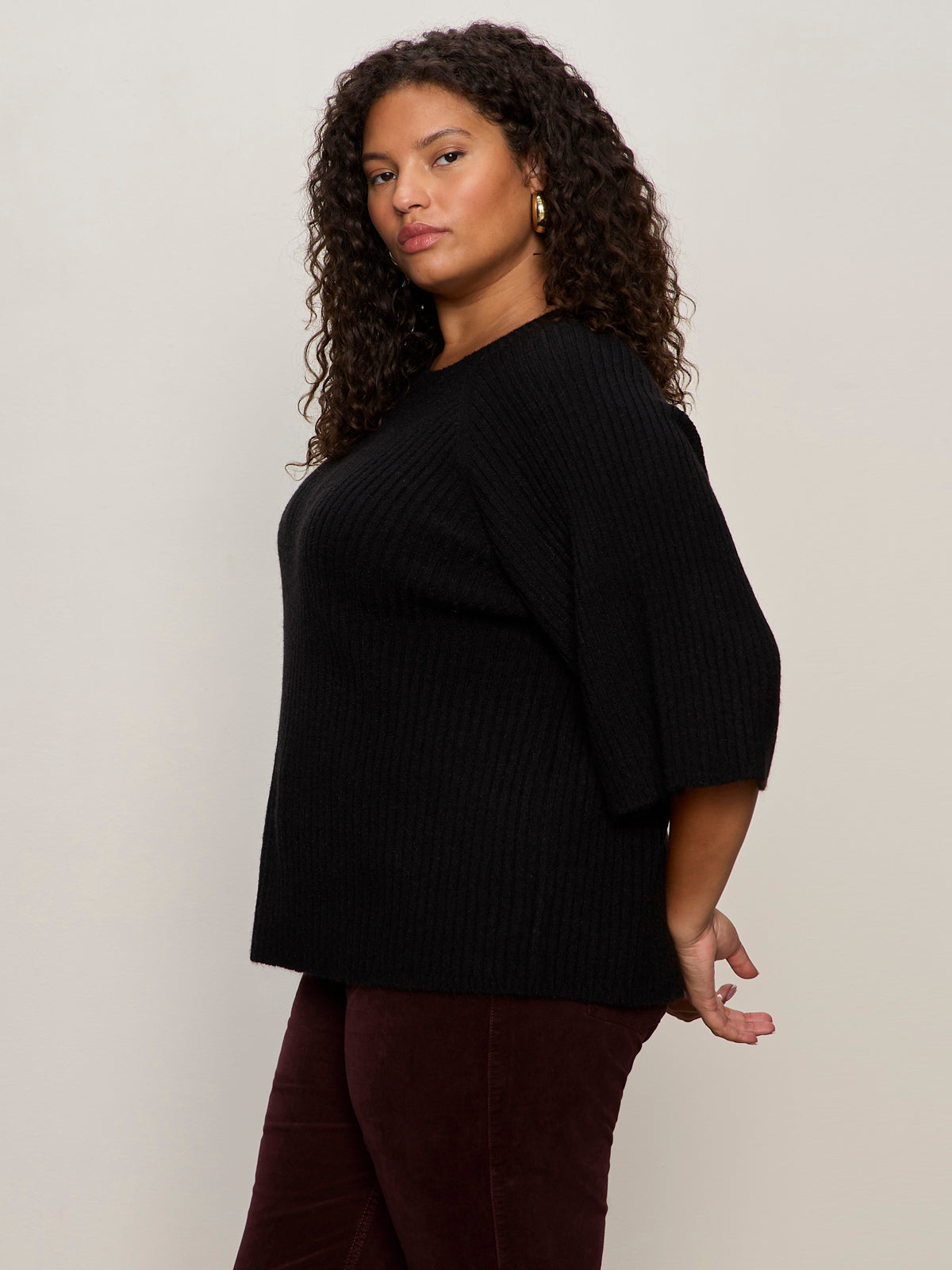 A woman with curly hair stands in profile against a light background, wearing Sanctuary Clothing's ribbed sweater tee in black (extended sizing) paired with burgundy pants.